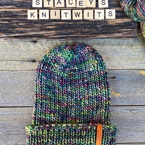 May include: A hand-knitted beanie in a vibrant mix of colors, including green, purple, and yellow. The hat features a folded brim and a small leather tag. The text "STACEYS KNITWITS" is spelled out in wooden letter tiles.