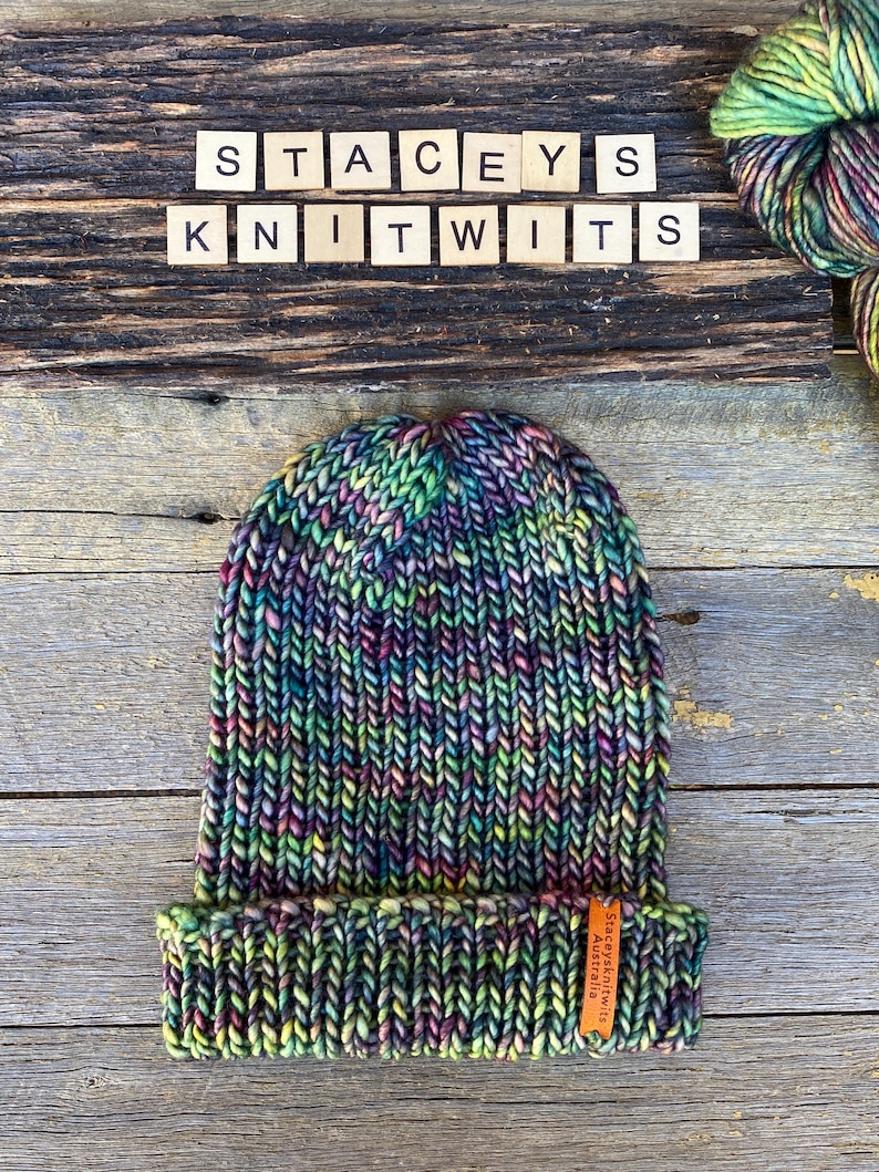 May include: A hand-knitted beanie in a vibrant mix of colors, including green, purple, and yellow. The hat features a folded brim and a small leather tag. The text "STACEYS KNITWITS" is spelled out in wooden letter tiles.