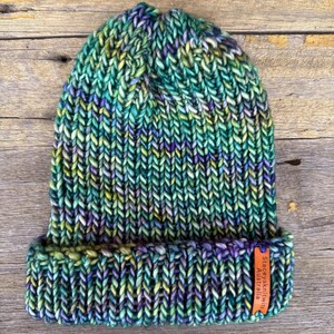 May include: A hand-knitted beanie in shades of green, blue, purple, and yellow. The hat has a folded brim and a tag that reads "Staceysknits Australia". The beanie is made of thick yarn and has a textured appearance.