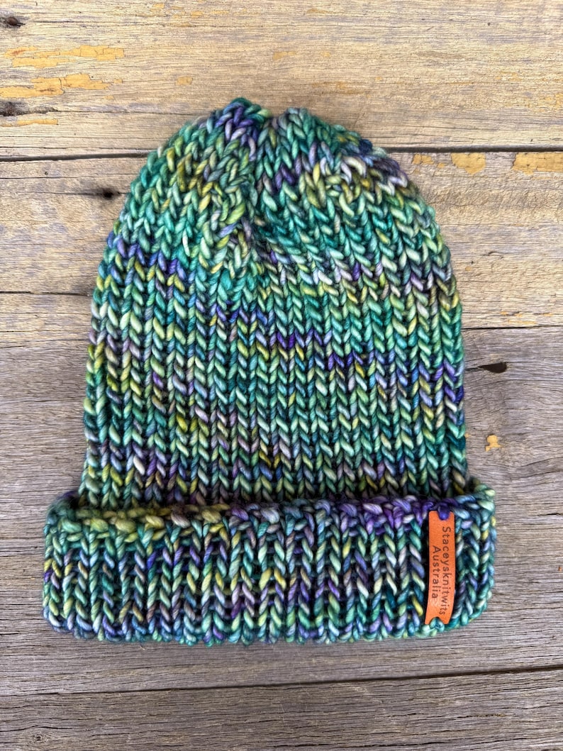 May include: A hand-knitted beanie in shades of green, blue, purple, and yellow. The hat has a folded brim and a tag that reads "Staceysknits Australia". The beanie is made of thick yarn and has a textured appearance.