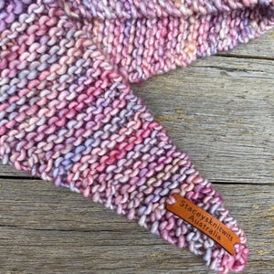 May include: A hand-knitted triangular scarf featuring a variegated yarn in shades of pink, purple, and soft blue. The textured garter stitch fabric includes a small, rectangular brown leather tag embossed with the text Staceysknitwits Australia.