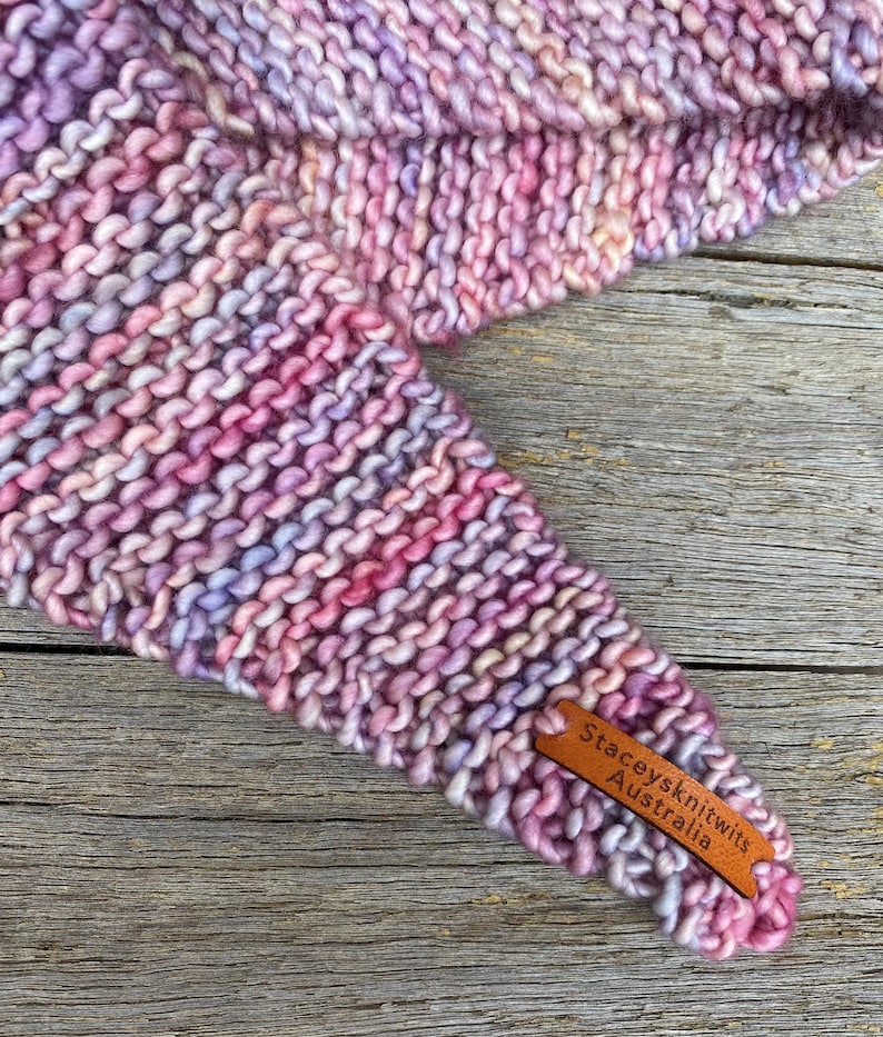 May include: A hand-knitted triangular scarf featuring a variegated yarn in shades of pink, purple, and soft blue. The textured garter stitch fabric includes a small, rectangular brown leather tag embossed with the text Staceysknitwits Australia.