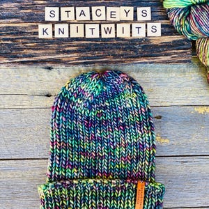 May include: A hand-knitted beanie in a vibrant mix of colors, including blue, green, pink, and yellow. The hat features a folded brim and a small leather tag. The text "STACEYS KNITWITS" is spelled out in wooden letter tiles above the hat.