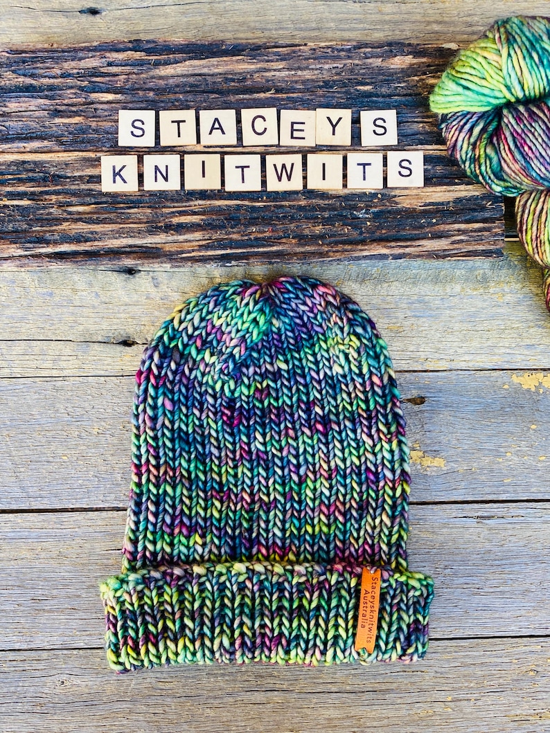 May include: A hand-knitted beanie in a vibrant mix of colors, including blue, green, pink, and yellow. The hat features a folded brim and a small leather tag. The text "STACEYS KNITWITS" is spelled out in wooden letter tiles above the hat.