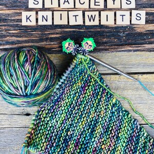 May include: A ball of variegated green, purple, and yellow yarn next to a partially knitted piece on a circular needle. Two small character-shaped stitch markers are attached to the needle. Wooden tiles spell STACEYS KNITWITS above the yarn.