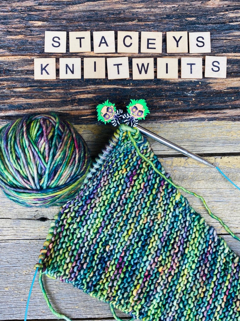 May include: A ball of variegated green, purple, and yellow yarn next to a partially knitted piece on a circular needle. Two small character-shaped stitch markers are attached to the needle. Wooden tiles spell STACEYS KNITWITS above the yarn.