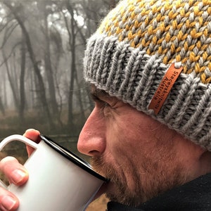 Può includere: Un uomo indossa un berretto di lana grigio e giallo con un'etichetta in pelle che recita "Stacey Australia" e beve da una tazza di smalto bianca.