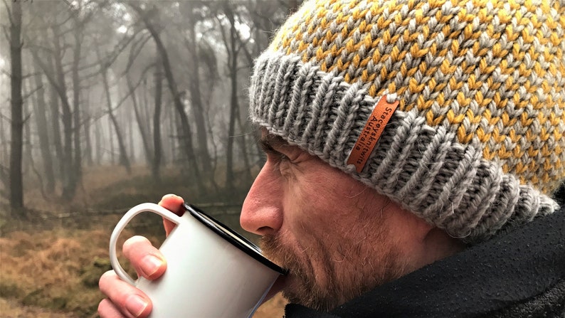 May include: A man wearing a gray and yellow knit beanie with a leather tag that reads "Stacey Australia" is drinking from a white enamel mug.