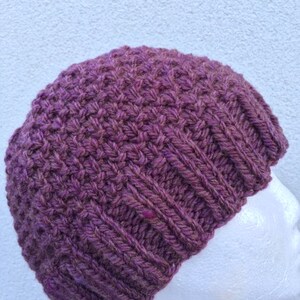 May include: A purple knitted beanie hat with a textured stitch pattern. The hat is shown on a white mannequin head.