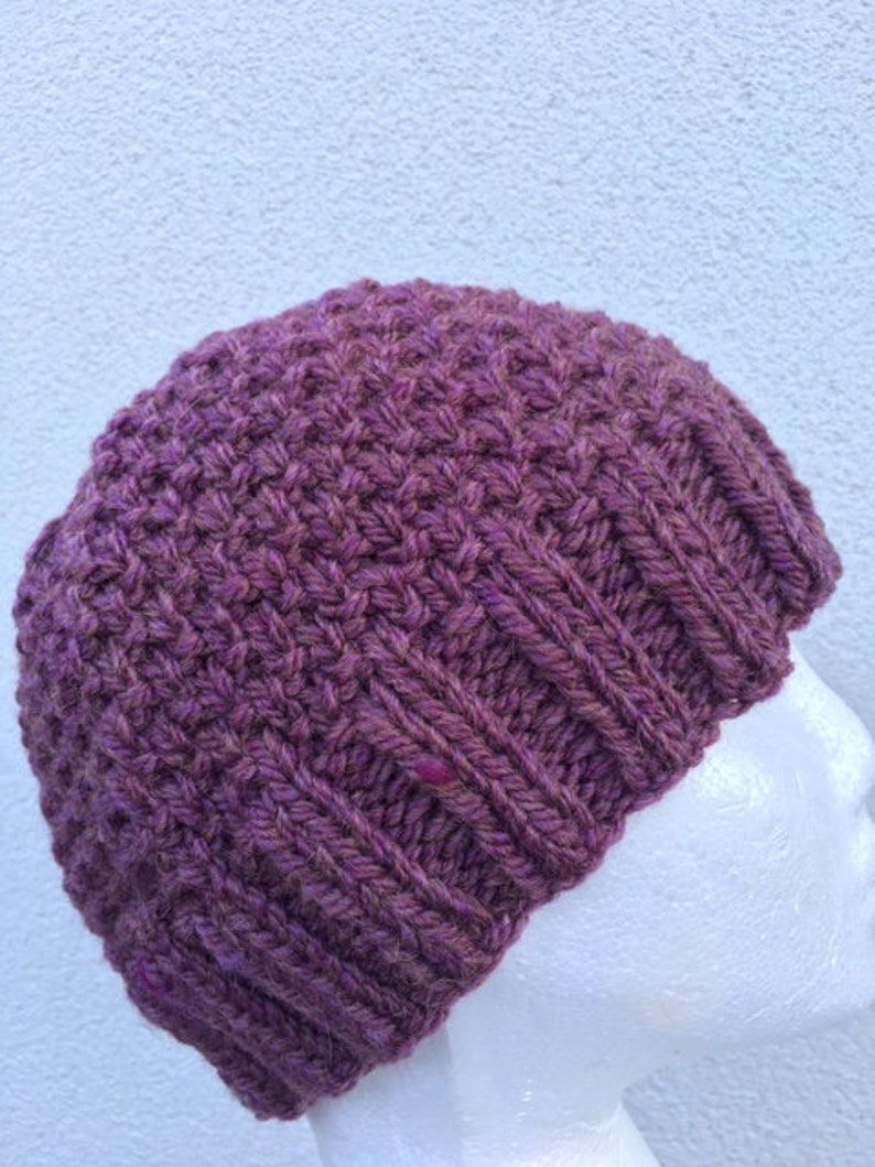 May include: A purple knitted beanie hat with a textured stitch pattern. The hat is shown on a white mannequin head.