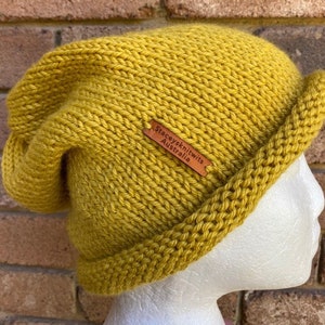 May include: A mustard yellow knitted beanie with a folded brim. The beanie has a brown leather label that reads "Stacey's Knitwits Australia".