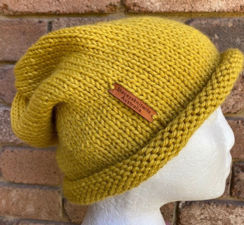 May include: A mustard yellow knitted beanie with a folded brim. The beanie has a brown leather label that reads "Stacey's Knitwits Australia".