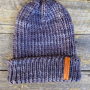 May include: A hand-knitted beanie in shades of purple and gray, with a ribbed design. The hat features a folded brim and a small, brown leather tag with the text "Staceysknitwits Australia". The hat is made of thick yarn.