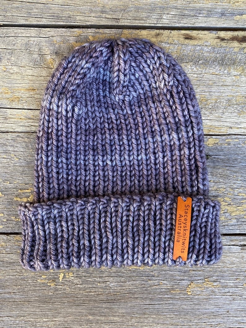 May include: A hand-knitted beanie in shades of purple and gray, with a ribbed design. The hat features a folded brim and a small, brown leather tag with the text "Staceysknitwits Australia". The hat is made of thick yarn.