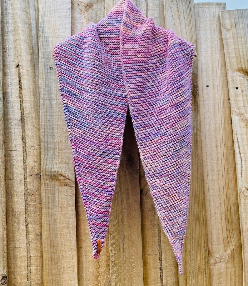 May include: A triangular knit scarf featuring a variegated color palette of pink, purple, blue, and orange yarn. The scarf is constructed with a garter stitch pattern and includes a small brown leather tag attached to one of the bottom points.