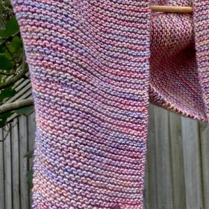 May include: A hand-knitted scarf featuring a garter stitch pattern with a variegated color palette of soft pink, lavender, and peach tones. The scarf is draped over a wooden rod, showcasing the textured knit construction and the blended yarn colors.