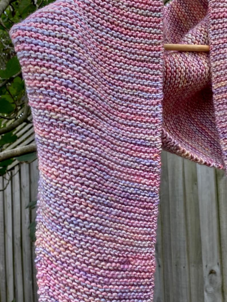 May include: A hand-knitted scarf featuring a garter stitch pattern with a variegated color palette of soft pink, lavender, and peach tones. The scarf is draped over a wooden rod, showcasing the textured knit construction and the blended yarn colors.