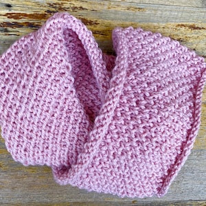 May include: A pink knitted infinity scarf. The scarf is made with a simple stitch pattern and has a soft, cozy feel.