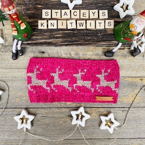 May include: A pink knitted headband with a reindeer pattern. The headband is on a wooden surface with white stars and a sign that says "Stacey's Knitwits".