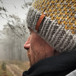 May include: A gray and yellow knit beanie with a brown leather label that reads "Adventure Awaits". The beanie is worn by a person standing in a foggy forest.