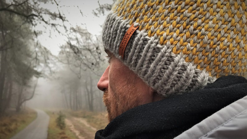 May include: A gray and yellow knit beanie with a brown leather label that reads "Adventure Awaits". The beanie is worn by a person standing in a foggy forest.