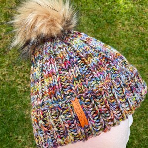 May include: Multicolored knitted beanie with a fluffy brown pom pom and a leather tag that reads "Stacey's Knits Australia".