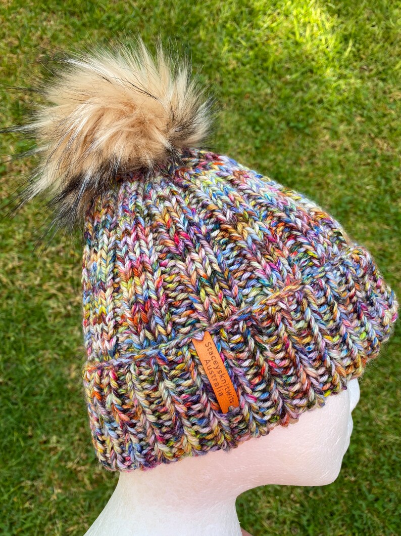 May include: Multicolored knitted beanie with a fluffy brown pom pom and a leather tag that reads "Stacey's Knits Australia".