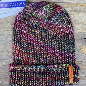 May include: A hand-knitted beanie hat in a vibrant mix of colors, including purple, pink, green, and yellow. The hat features a folded brim and a textured knit pattern. The label reads "Staceysknitwits Australia".