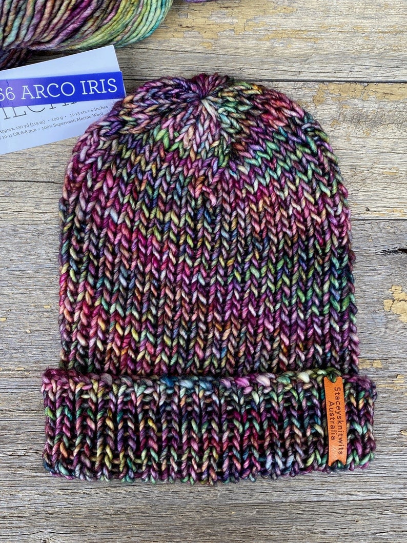 May include: A hand-knitted beanie hat in a vibrant mix of colors, including purple, pink, green, and yellow. The hat features a folded brim and a textured knit pattern. The label reads "Staceysknitwits Australia".