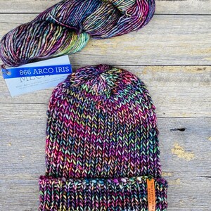 May include: A hand-knitted beanie in a vibrant rainbow of colors, with a folded brim. A skein of matching yarn sits above the hat. The yarn label reads "866 Arco Iris". The hat has a small tag that reads "Staceykniits Australia".