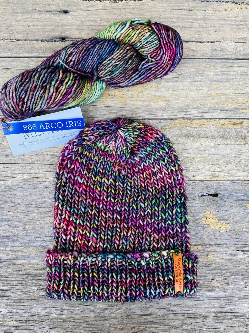 May include: A hand-knitted beanie in a vibrant rainbow of colors, with a folded brim. A skein of matching yarn sits above the hat. The yarn label reads "866 Arco Iris". The hat has a small tag that reads "Staceykniits Australia".