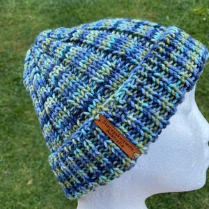 May include: Blue, green, and white knitted beanie with a brown leather tag that reads "Staceyknits Australia".