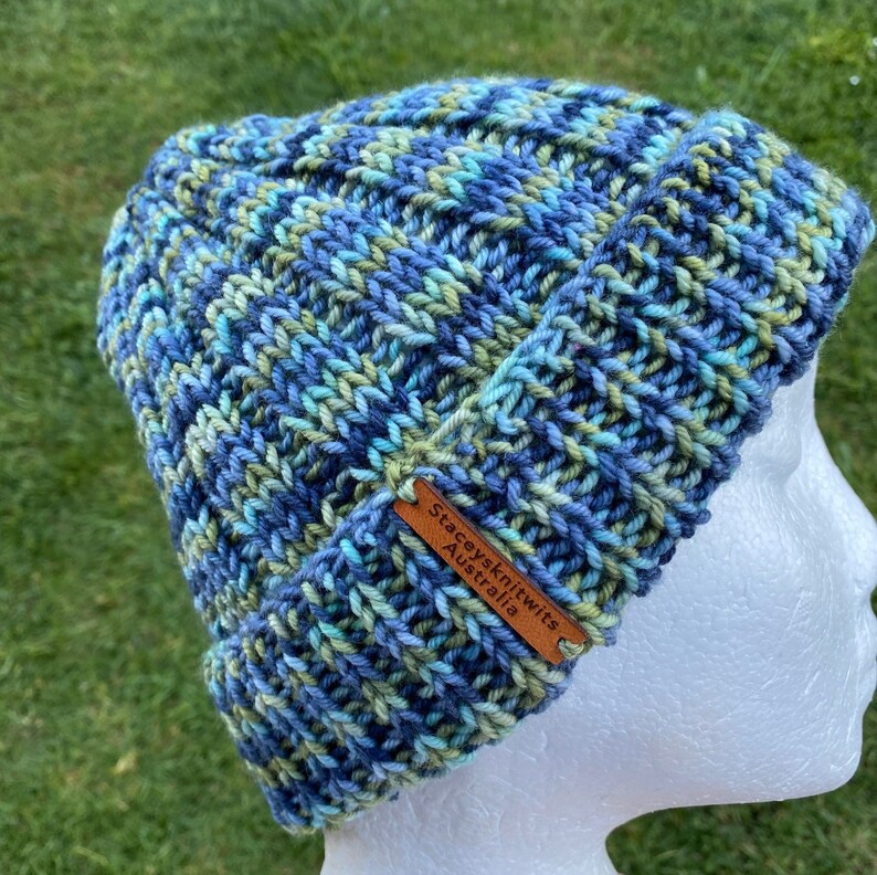 May include: Blue, green, and white knitted beanie with a brown leather tag that reads "Staceyknits Australia".