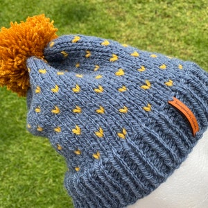 May include: A blue knitted beanie with a yellow pom pom and yellow heart pattern. The beanie has a brown leather tag with the text "Exceptional Designs" on it.