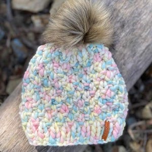 May include: A colorful knitted beanie with a fluffy pom pom on top. The beanie is made with a chunky yarn in shades of pink, blue, yellow, and white.