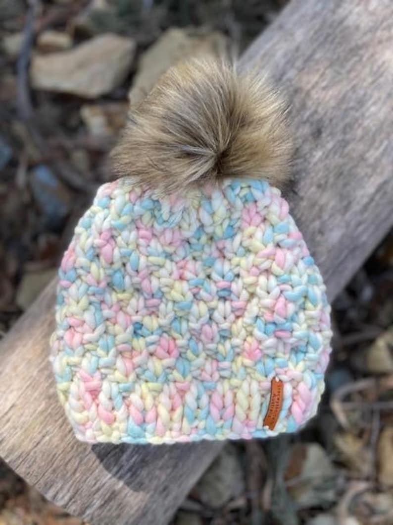 May include: A colorful knitted beanie with a fluffy pom pom on top. The beanie is made with a chunky yarn in shades of pink, blue, yellow, and white.