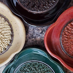 Peut inclure: Quatre assiettes en céramique avec des motifs et des couleurs différents. Les assiettes sont toutes rondes et ont un bord surélevé. La première assiette est beige avec un motif brun et blanc. La deuxième assiette est noire avec un motif brun. La troisième assiette est rouge avec un motif brun. La quatrième assiette est verte avec un motif noir.