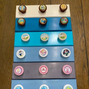 May include: A set of 10 white drawer knobs with colorful cartoon animal and character designs. The knobs are arranged in rows on a set of painted wooden drawer fronts. The drawer fronts are painted in various colors, including white, blue, turquoise, gray, and pink.