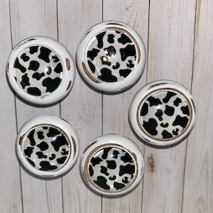 May include: Five white round knobs with a black and white cow print design. The knobs have a distressed white finish and a brown ring around the edge.