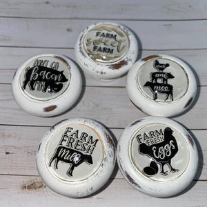 May include: Five white ceramic knobs with black and white farm-themed designs. The designs include a pig, a cow, a chicken, and text that says "Don't Go Bacon", "Farm Sweet Farm", "Farm Fresh Milk", "Cluck Ank Moo", and "Farm Fresh Eggs".