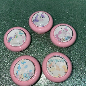 May include: Five pink round buttons with a unicorn design. Each button has a different unicorn design and text. The text on the buttons reads "unicorn", "dreams", "magical", and "unicorn".
