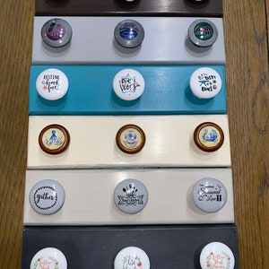 May include: A set of 10 decorative cabinet knobs with various designs and sayings. The knobs are white with black lettering and are arranged in rows on a wooden surface. The sayings include "Farm Fresh", "Resting Beach Face", "Beach Vibes", "Better at the Beach", "Gather", "Nana's Kitchen", "The Kitchen Seasoned with Love", "Follow That Dream", "Live Simply", and "Better Than Ever".