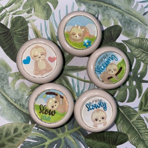 May include: Set of five round, beige knobs with sloth designs. Each knob features a different sloth with a unique phrase, including "Slow Down", "I'd Rather Be Sleeping", and "Go Slowly".
