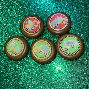 May include: Five wooden knobs with different construction vehicle designs. Each knob features a yellow vehicle with black wheels on a colorful background. The knobs are brown with a yellow and black border.