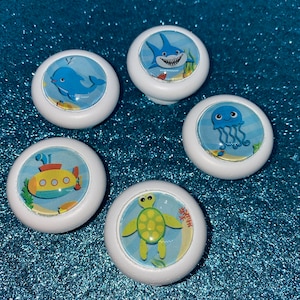 May include: Five white round knobs with different ocean-themed designs. The designs include a shark, a whale, a jellyfish, a submarine, and a sea turtle.