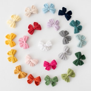Könnte beinhalten: Eine Ansammlung farbenfroher, gehäkelter Schleifen in verschiedenen Farbtönen wie Gelb, Rot, Rosa und Blau, angeordnet auf einer weißen Oberfläche. Die handgefertigten Schleifen haben eine strukturierte Optik, die auf ein Bastel- oder DIY-Thema hindeutet.