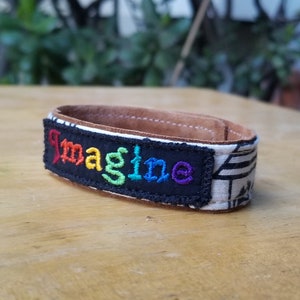 May include: A brown leather bracelet with a black fabric patch that says "Imagine" in rainbow colors.