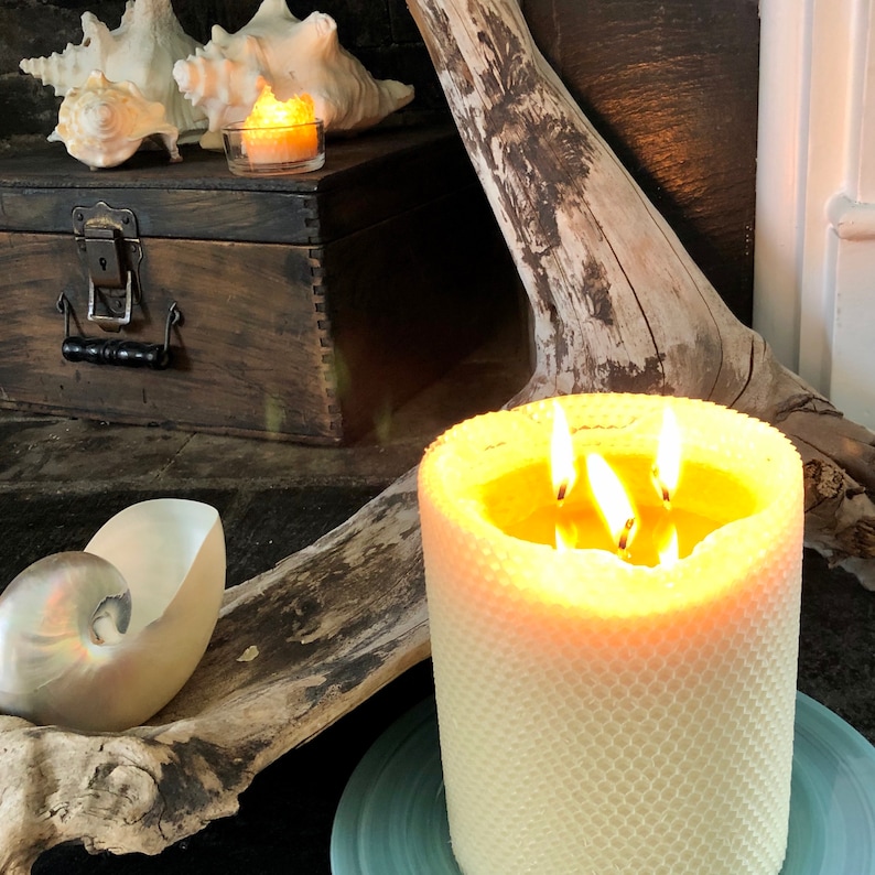 Op de afbeelding: Een witte honingraat-kaars met drie lonten die branden, op een blauw en wit bord. De kaars staat op een houten oppervlak met schelpen en een kleine houten doos.