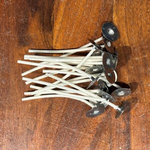 May include: A collection of white candle wicks, each featuring a circular metal base at one end. The wicks are arranged in a loose cluster, showing the contrast between the white fiber material and the metallic silver-toned circular supports.