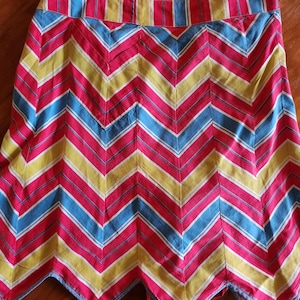 May include: A colorful skirt with a chevron pattern. The skirt features alternating stripes of red, yellow, blue, and white. The hemline has a scalloped edge. The skirt is made of a lightweight fabric.